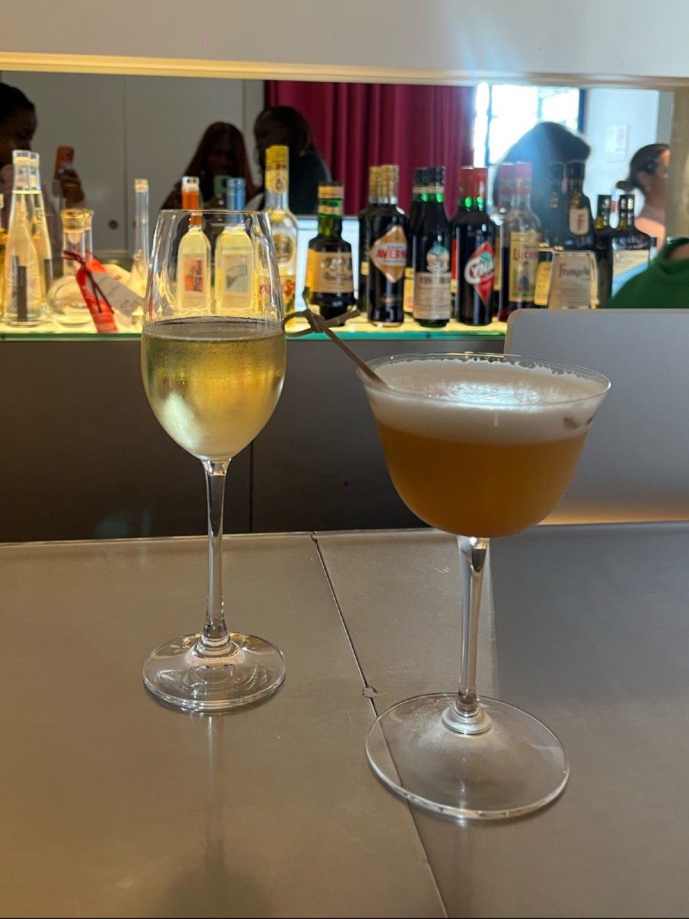 A glass of white wine and an amber cocktail on the counter in front of a lit back bar of bottles