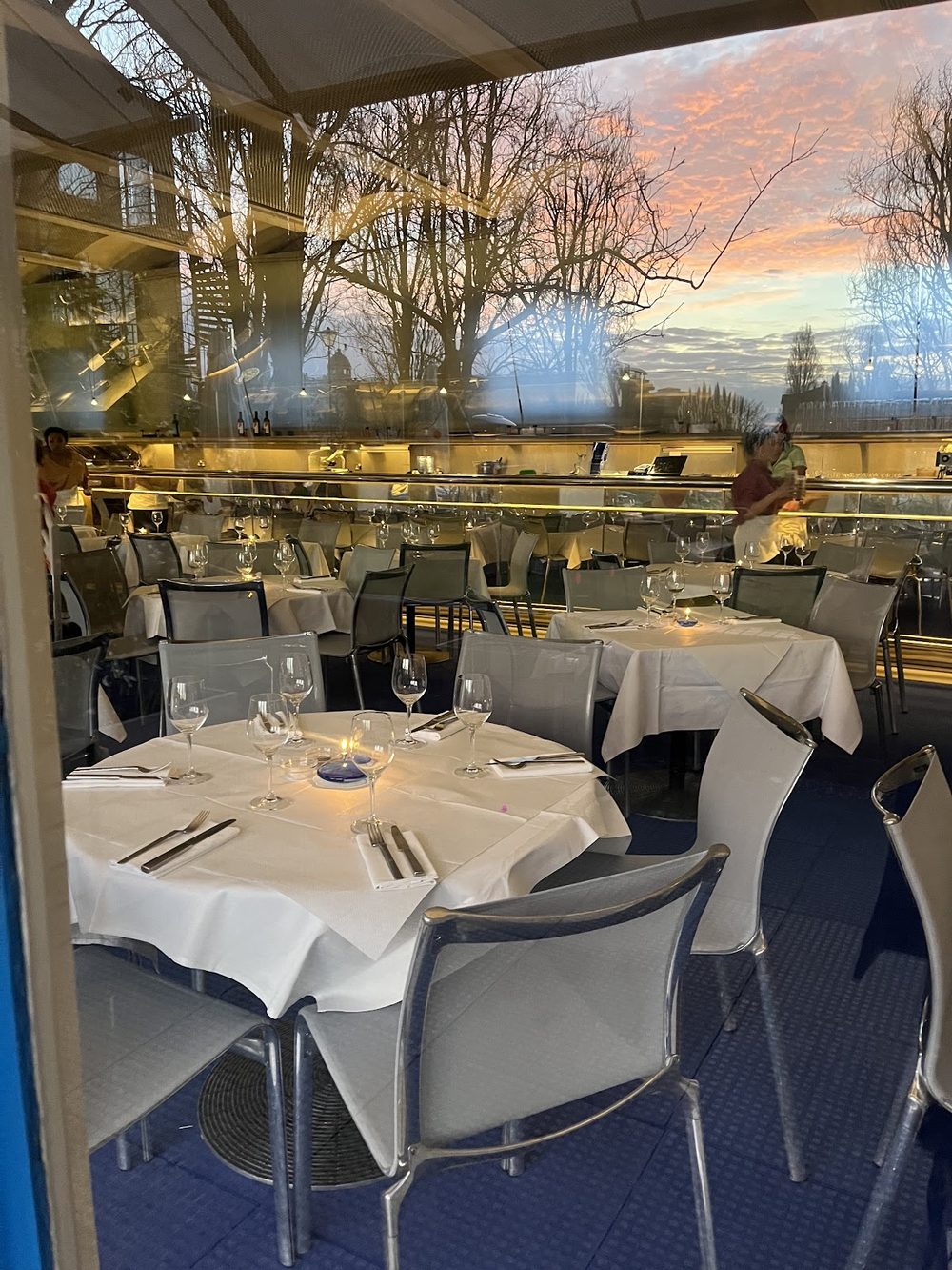 White-clothed tables set in front of a full-height window with a pink sunset over the trees outside